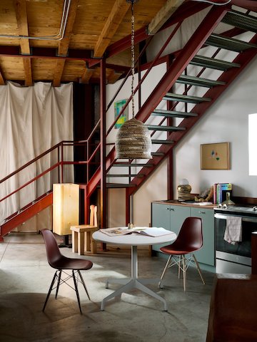 An industrial-style double-height ceiling space with dark red metal open staircase and a small galley kitchen featuring a small white Eames Table and two brick red Eames Shell Chairs with dowel legs.