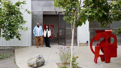 Benjamin Critton and Heidi Korsavong—framed between small trees— stand outside of Marta gallery, an industrial indoor-outdoor auto repair garage with white metal siding and a cement walkway.