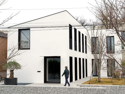 An outside view of Naoto Fukasawa walking into his studio in Tokyo, with stark white walls and black-framed windows and a neatly shaped rock wall.