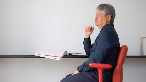 Designer Naoto Fukasawa sits in profile in a red Asari Chair by Herman Miller at a minimal desk with an open magazine laid in front of him.