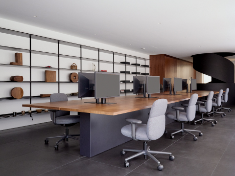 A line of five light gray Asari Chairs by Herman MILLER sit at a long wooden desk system with black computer monitors. Behind the desk is a minimal black shelving unit with various wooden sculptures placed and a modern black spiral staircase.