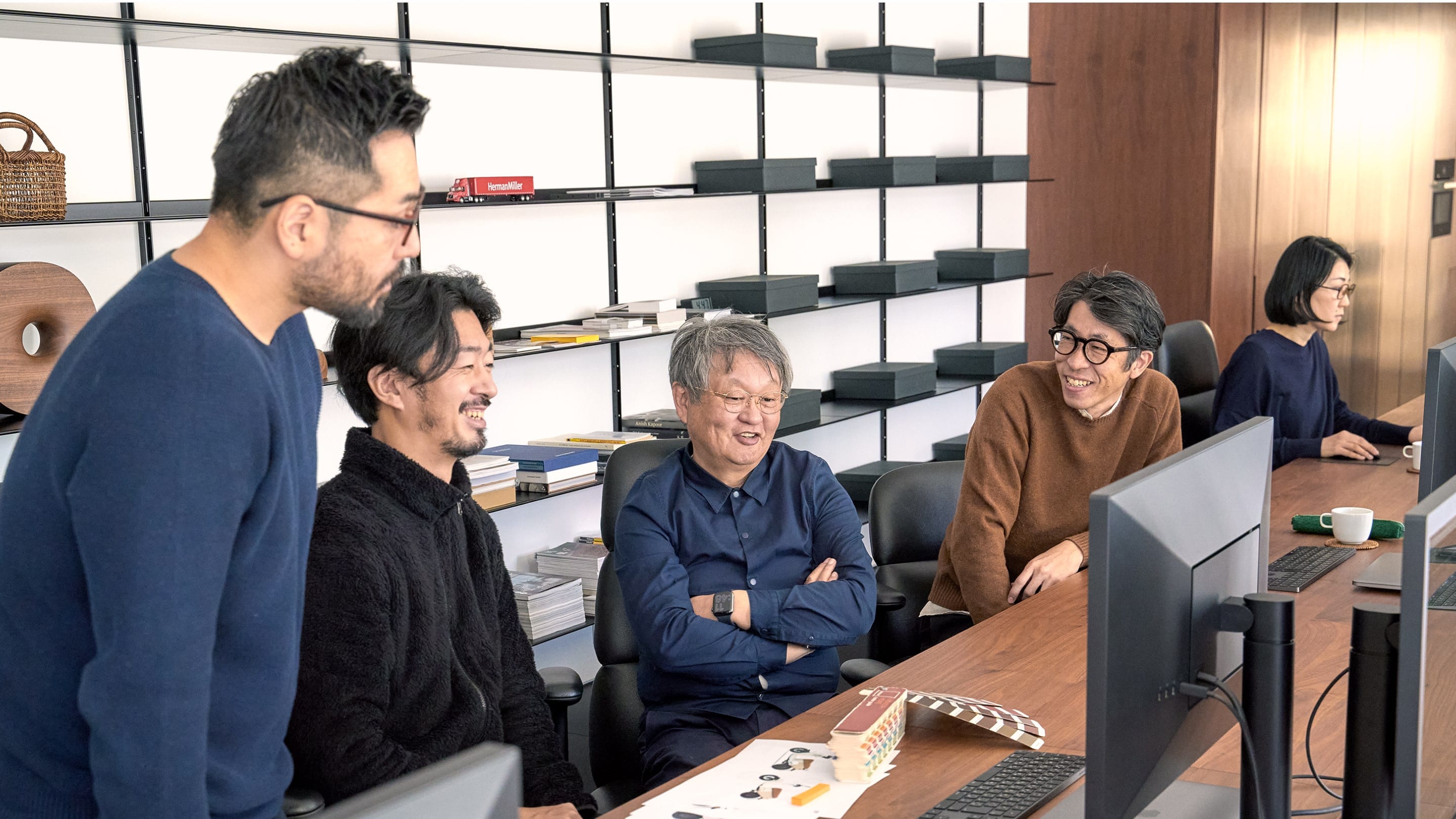 Naoto Fukasawa sits in the center of a group of five designers at a long wooden desk, with a minimal black shelf behind them featuring wooden sculptures, various ephemera, and black file boxes. In front of them are a Pantone swatch deck and computer monitors.