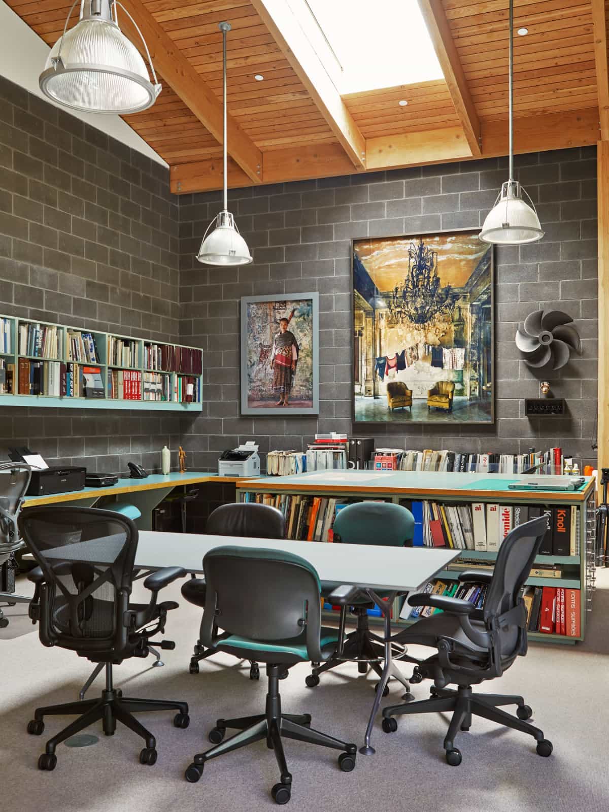 Don Chadwick's industrial-style LA studio with dark brick walls and wood paneled ceiling. Featured are a small conference table and several dark gray and black Aeron and Equa Chairs, and overstuffed bookshelves nearby.