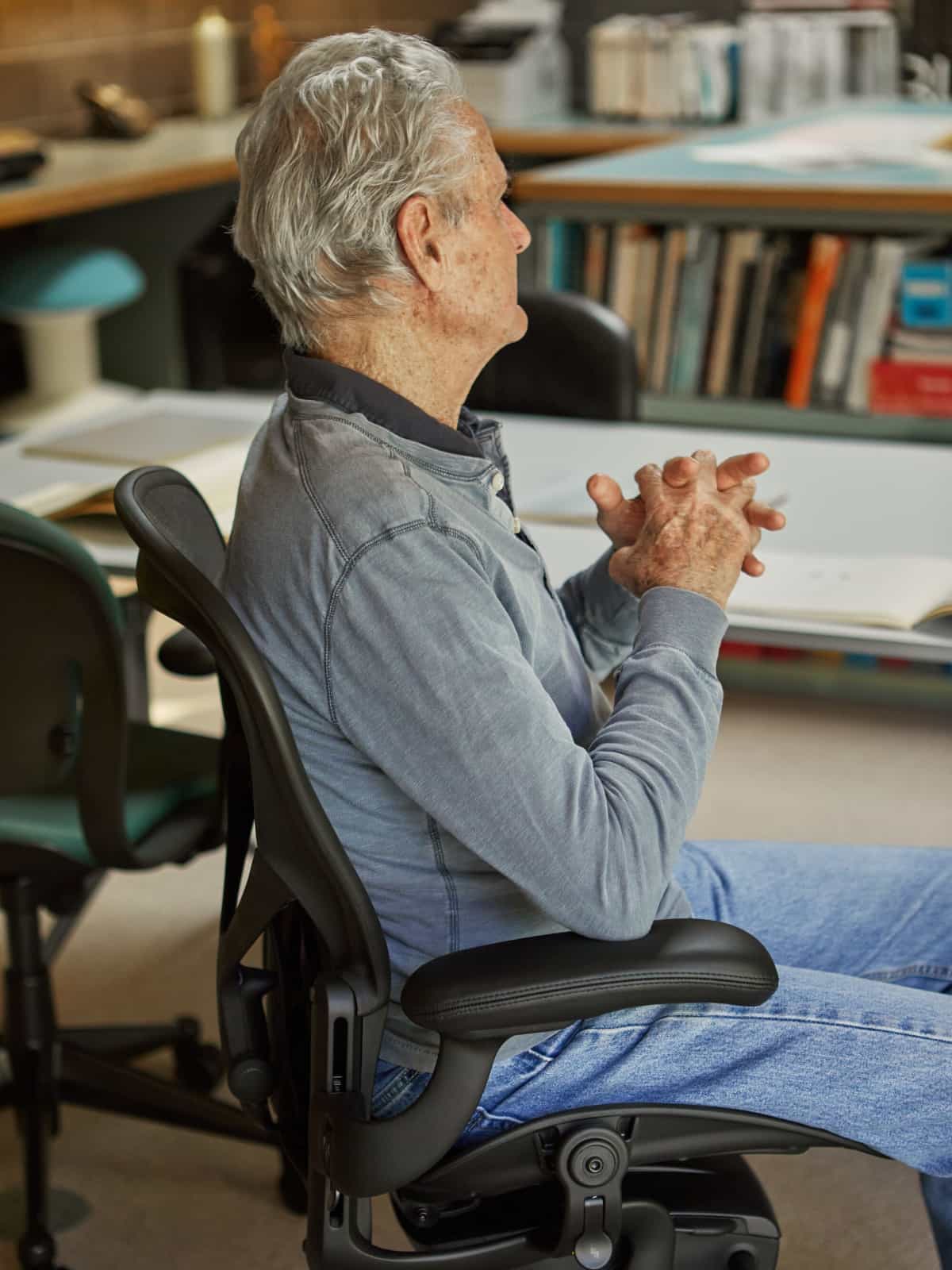 A side view of Don Chadwick sitting in the re-engineered Aeron Chair in Onyx Ultra Matte color.