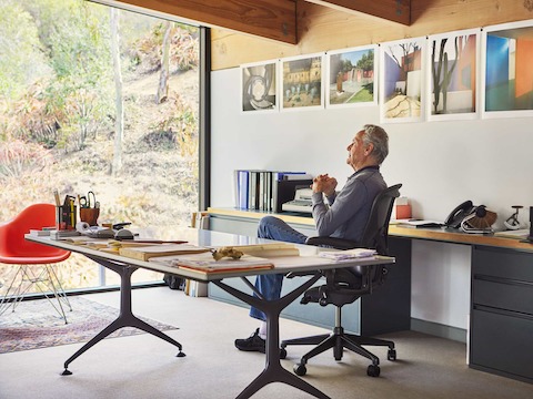Don Chadwick looks out a window while sitting in his original dark gray Aeron Chair at a long, organized desk with storage behind him.