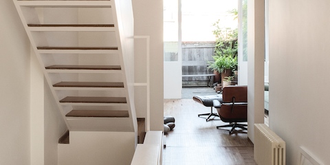 The London home of designer Michael Anastassiades, featuring a white staircase and a black leather and walnut Eames Lounge Chair facing a doorway to a patio with plants.  
