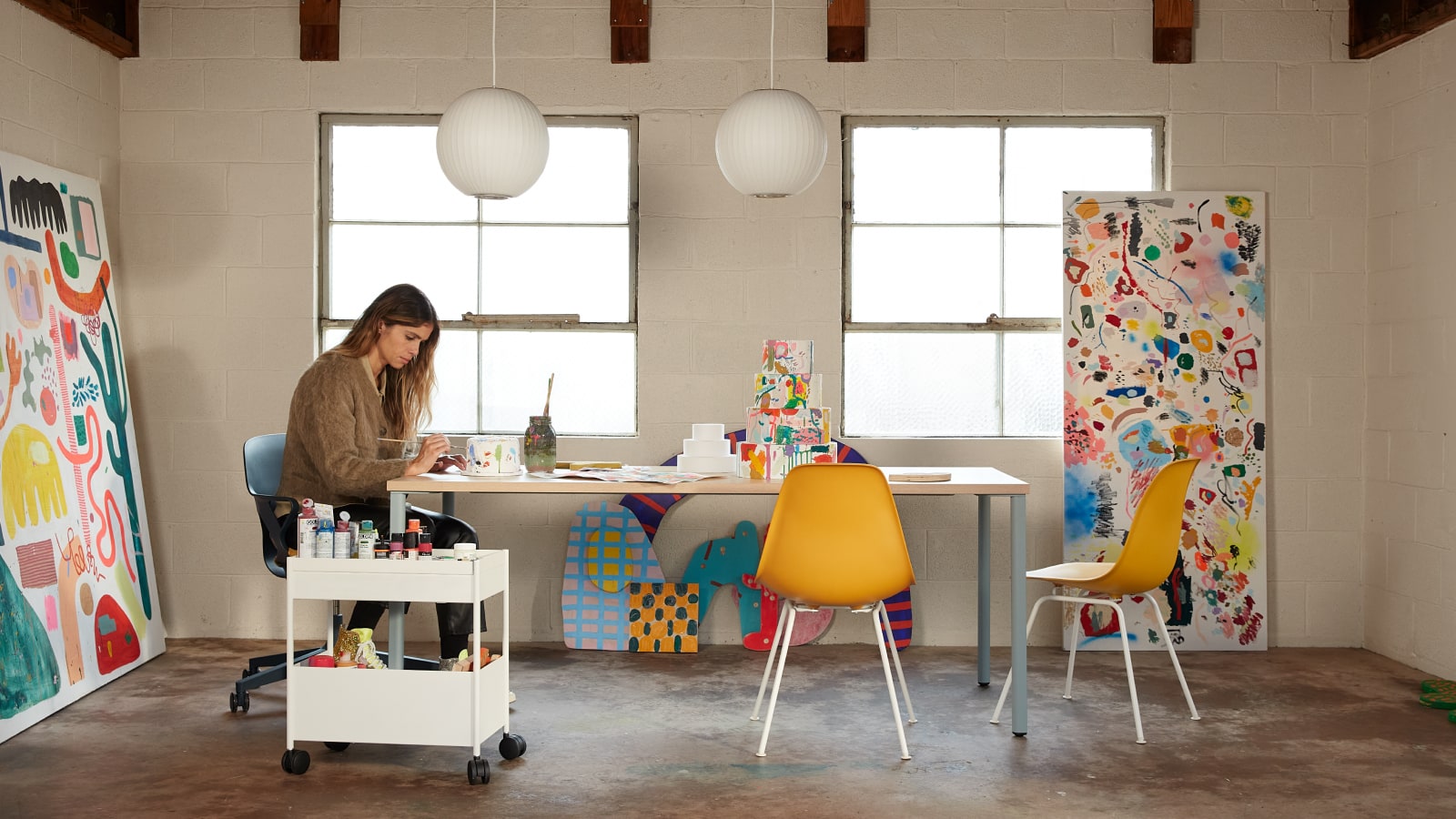 L'artista Kindah Khalidy siede a un tavolo su una seduta Zeph; nell'ambiente, due sedute monoscocca Eames gialle.