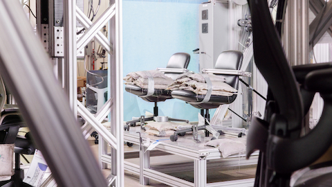 Two black work chairs sit with gray weighted bags in their seats, strapped to a testing machine.