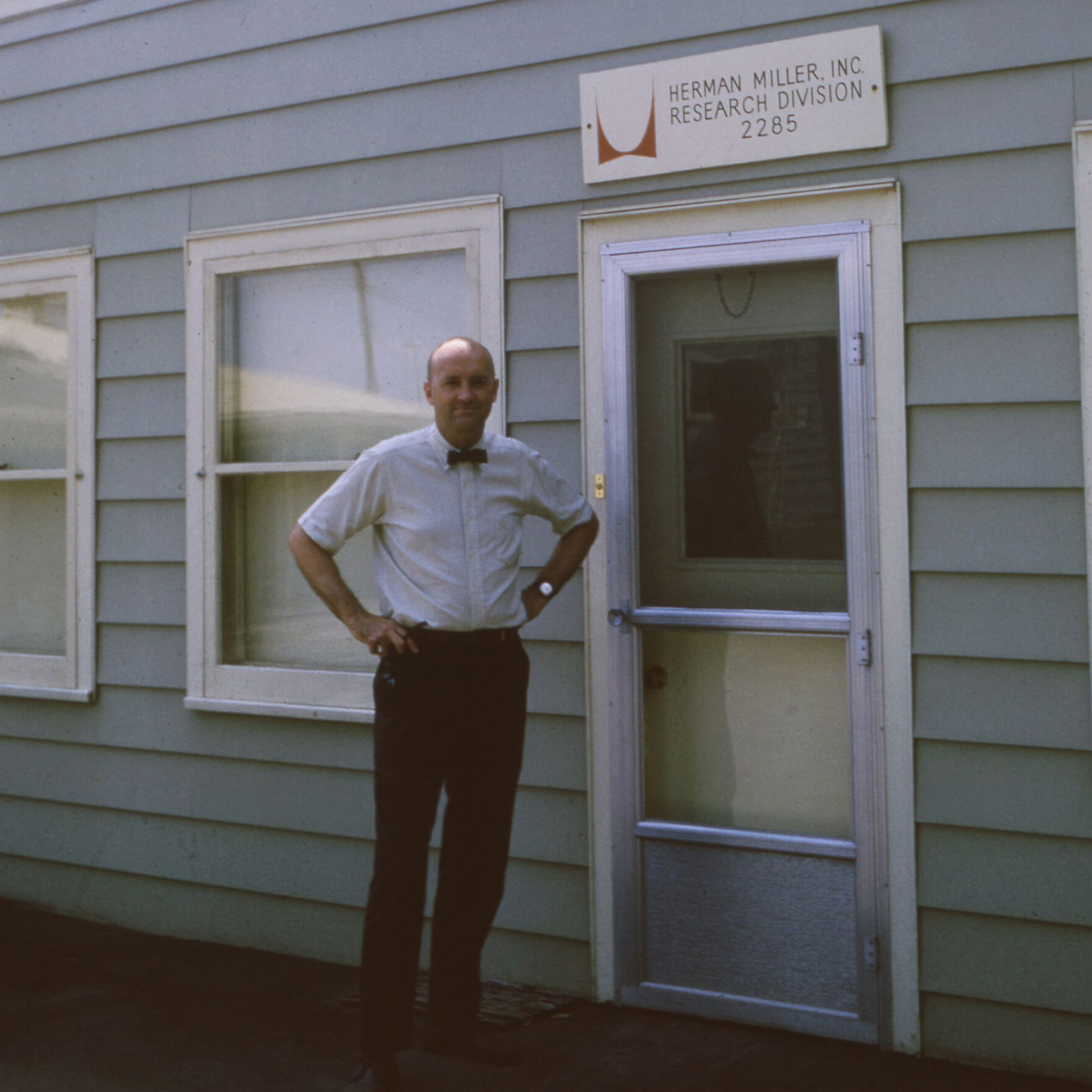 Propst standing outside in front of a building with light blue siding, two windows with white frames, a screen door with white frame, and white sign with Herman Miller red logo and black lettering that reads 'Herman Miller, Inc. Research Division, 2285.'