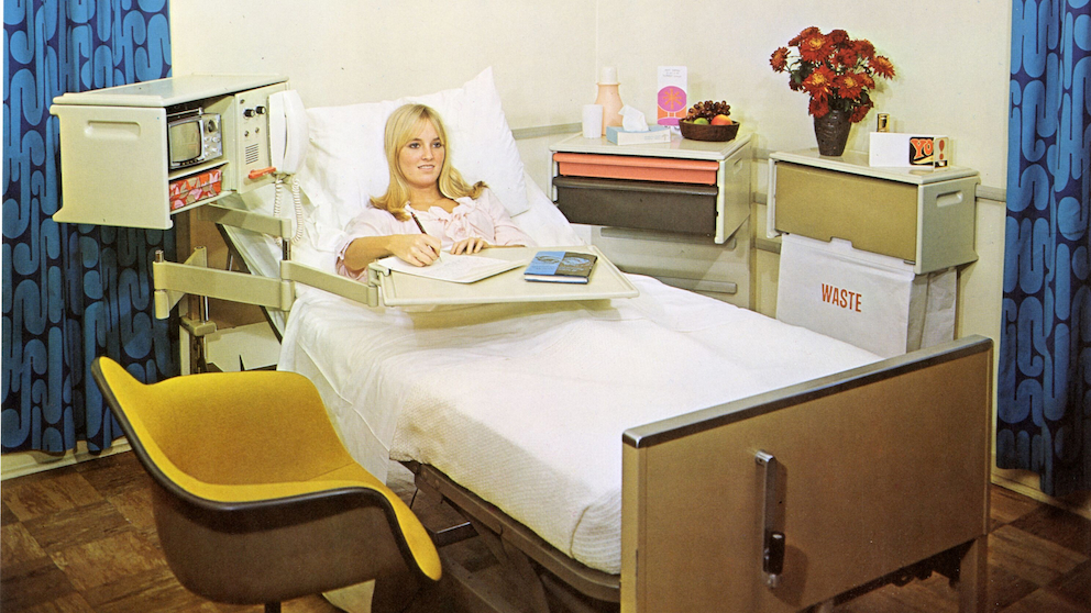 Woman in hospital bed sitting up and writing. On the left, yellow Eames molded plastic chair. On the right, Co/Struc overbed arm tray, arm-mounted nurses’ call, trimline phone, solid-state TV, and patient sub-shelf. Waste bag on wire hanger. L cart with storage drawers. Blue curtains in the background.