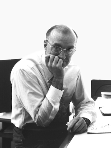 Archival black and white portrait of Robert Propst, seated at a desk leaning forward with the palm of his hand over mouth. 