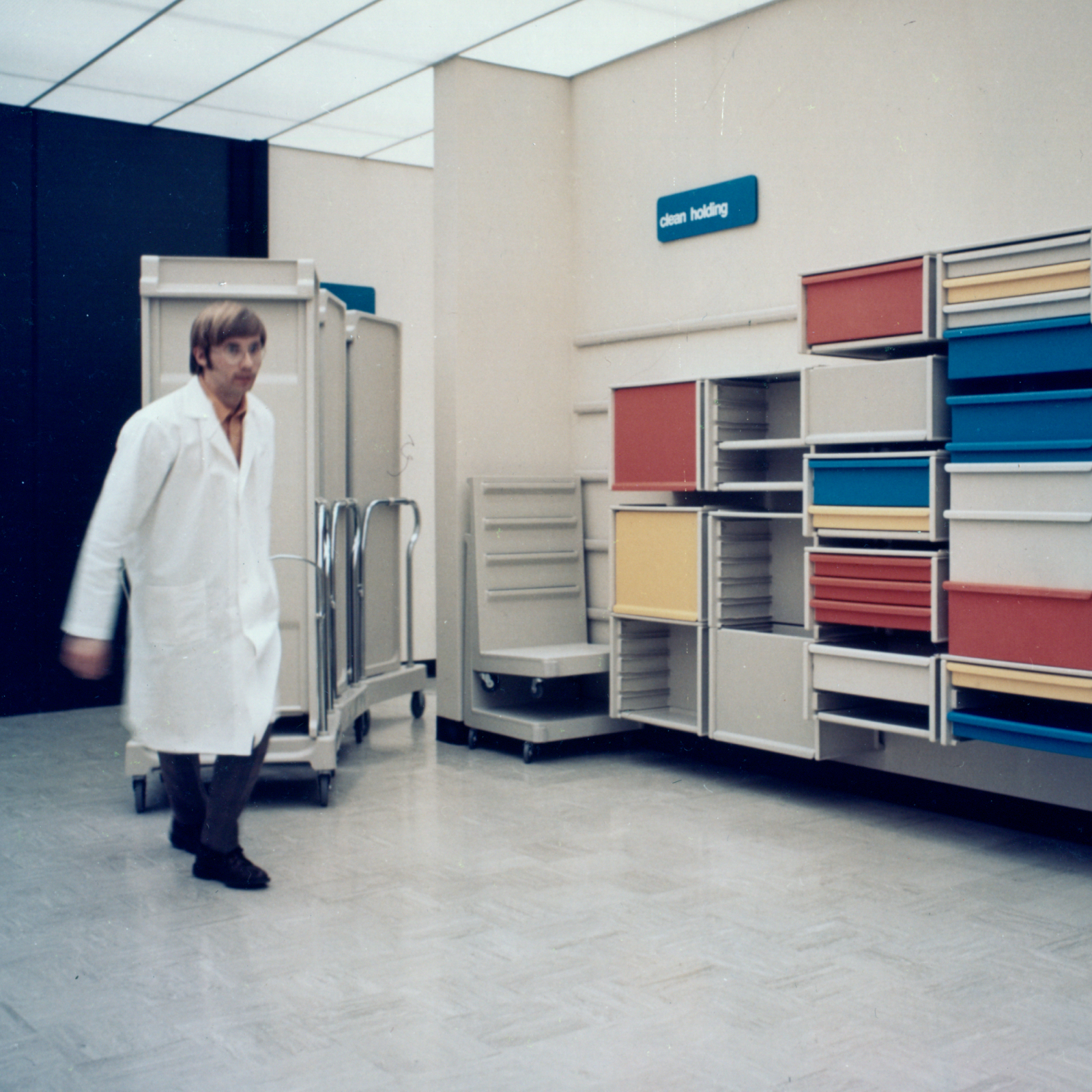 Archival photo of a man in white lab coat pulling Co/Struc lockers on transport cart in motion. Co/Struc wall-mounted open lockers with drawers in red, yellow, blue, and white background. Blue sign on the wall with white lettering that says 'clean holding.'