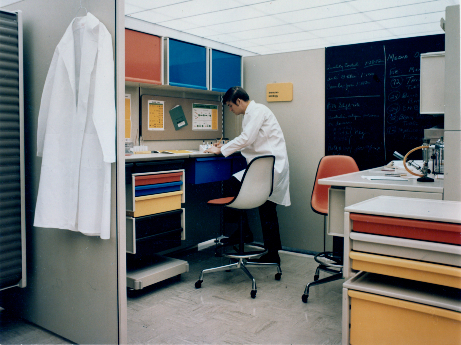 Lab room with a clinician in white coat, white coat hanging, Co/Struc system with overhead lateral storage, wall- and cart-mounted storage, and drawers in red, blue, and yellow. Two red stools with white back and black frame.