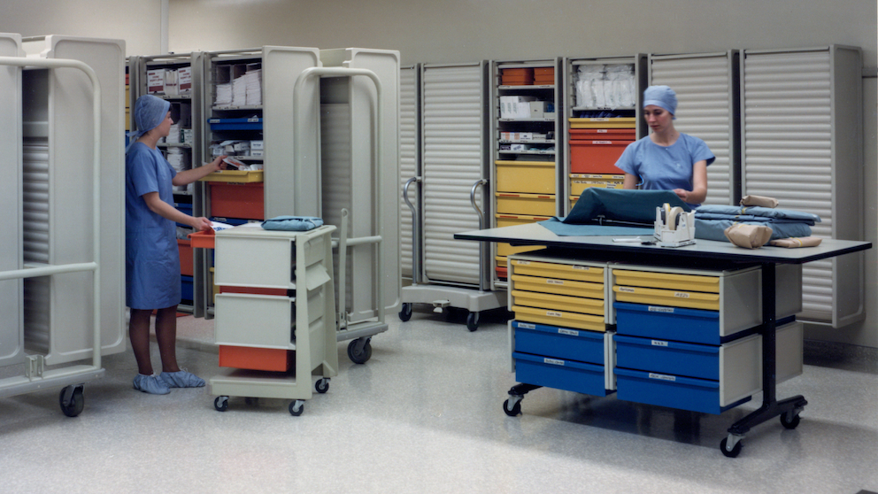 Two members of clinical staff in blue scrubs in a hospital storage room with Co/Struc System L cart with orange drawers, open and closed wall-mounted lockers with yellow and orange drawers, lockers on transport carts, table with wheels, storage with yellow and blue cabinets underneath.