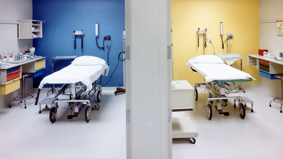 Mirrored exam rooms, left wall blue, right wall yellow. Co/Struc system nurses station with surface, wall- and cart-mounted storage, and drawers. Exam room supplies and seating.