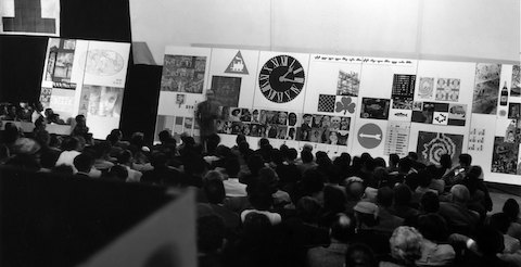 Una imagen en blanco y negro de un hombre hablando ante una multitud sentada.