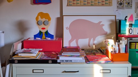 Stacks of books, notes, artist supplies and colourful art on top of a grey flat-file cabinet.