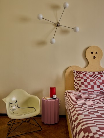 A cream-coloured Eames Moulded Fibreglass Armchair with a Saul Steinberg cat design next to a table and bed in Olimpia Zagnoli's home.