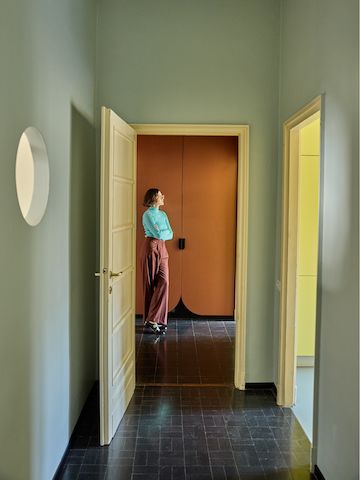 Olimpia Zagnoli standing in her home at the end of a hallway, just beyond an open door.