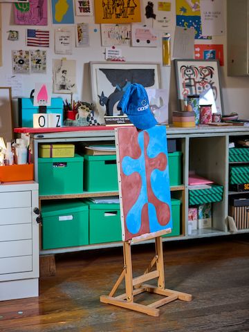 A blue and brown abstract painting on an easel in Olimpia Zagnoli's studio, in front of shelves of green boxes and a wall filled with small pieces of art.