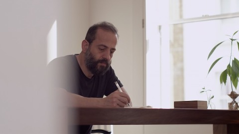 Designer Michael Anastassiades Zeichnung an einem Tisch. Wählen Sie, um zu einem Artikel und Video über Anastassiades zu gelangen.
