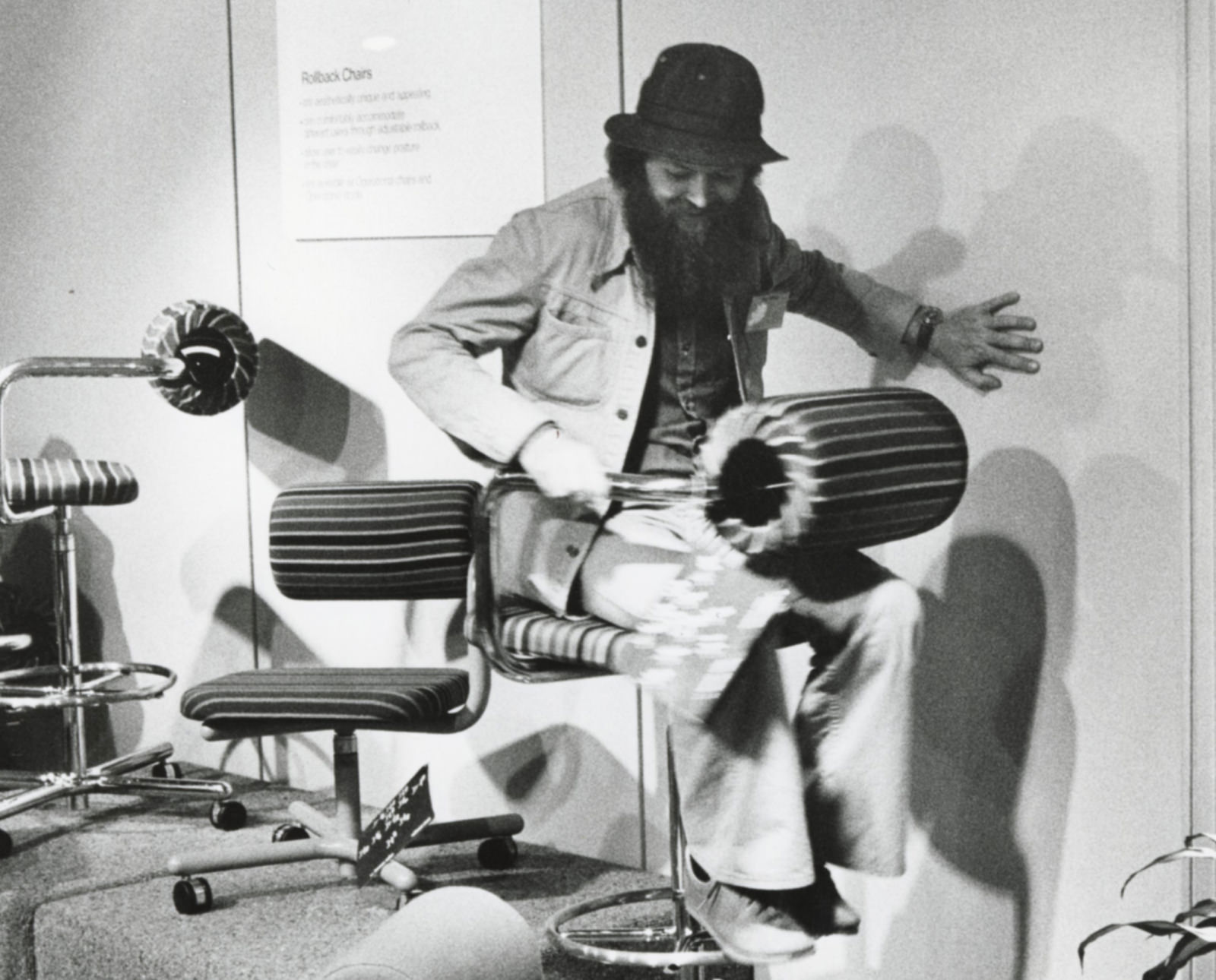 Ray Wilkes seated backwards on stool-height Rollback chair on raised platform surrounded by various chair models during its debut at Neocon in 1977.