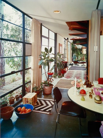 This color photo is a view of the kitchen and into the living room. A kitchen table with colorful flowers and objects is in the foreground. A white rug with pink diamonds is on the floor in the background.