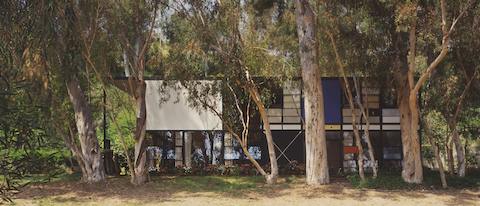 This color photos is a view of the Eames House from the front, showing off white, blue, and red panels. Eucalyptus trees and flowers line the front of the house.
