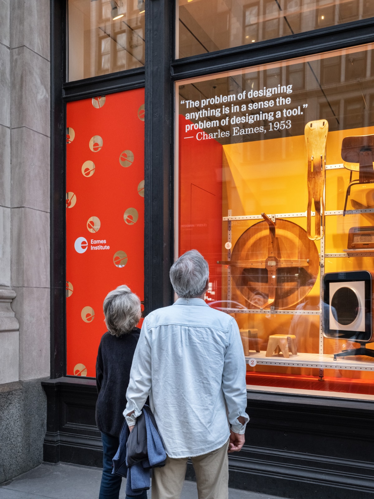 Two passerby look at a streetfront window featuring a plywood leg splint, a plywood coffee table, and a midcentury-style floor speaker.