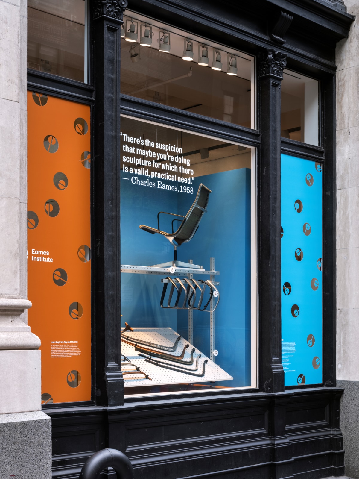 A window display in a large, granite building features a blue- and orange- exhibit showing chair components in wood and aluminum underneath a finished chair in aluminum with a mesh seat.