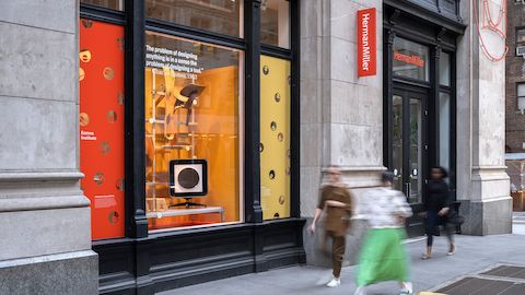People walk on a sidewalk in front of a Herman Miller store whose window is decked out with a red and orange exhibition setting housing a range of plywood furniture and décor. 